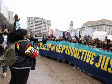 A woman speaks into a microphone, addressing a crowd behind a banner reading "Rise up for Reproductive Justice" at a January 22 protest in Washington, DC.