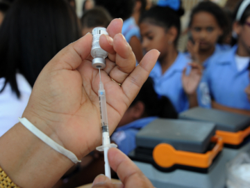 Close-up of a syringe held by a health ministry employee preparing to vaccinate girls against the HPV virus with several girls wearing sky blue uniform shirts in a blurred-out background.