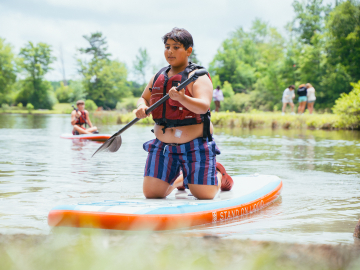 A boy in striped shorts and a life vest, with an insulin patch on his abdomen, paddles a canoe at Camp Ho Mita Koda