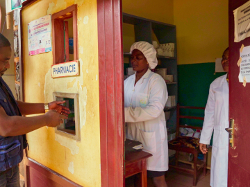 A man wearing a blue vest puts his hands out to receive medicine through a window cut out in a yellow-painted pharmacy.