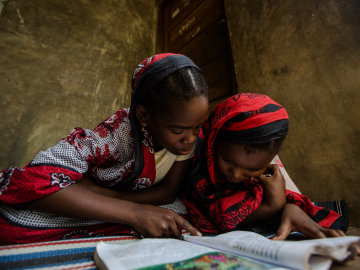 Two girls dressed in red and black garments sit on the floor reading USAID malaria educational materials in Zanzibar, Tanzania.