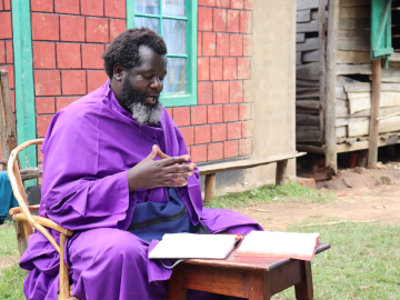 Eliud Wekesa, hands clasped, with a graying beard and wearing a bright purple robe, speaks to visitors outside his home and church compound in Tongaren, Bungoma County, Kenya, on February 29, 2024.