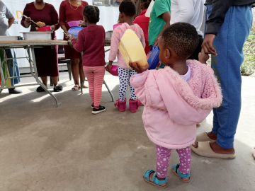 Children, including a toddler in a pink coat holding a yellow lunch container, line up to collect a meal.