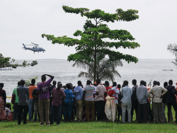 Dozens of bystanders look on as they watch rescuers in a helicopter search the site of a capsized cruise boat on Lake Victoria near Mutima village, south of Kampala, Uganda, on a grey, cloudy day.
