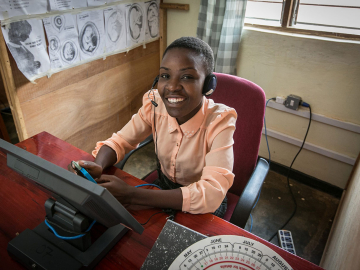A health care worker wearing a peach button-down blouse sits at a desk wearing a headset to connect with hotline callers in Lilongwe, Malawi.