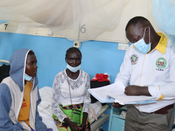 A doctor, standing and looking over a chart, attends to a patient seated on a cot.