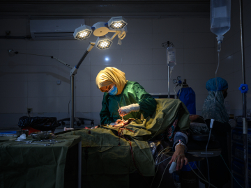 A female surgeon wearing a blue surgical mask and a yellow headscarf operating on a patient.