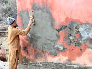 Mohammad Aftab, a man of about 30 years old with a blackish-brown beard, wearing a tan tunic top and pans and a blue and white hat, works applying a grayish, concrete-like mixture to a pink wall.