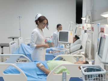 A nurse wearing a white uniform and hat stands next to a patient lying in a hospital bed and wearing a pastel green gown.