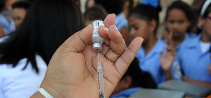 Close-up of a syringe held by a health ministry employee preparing to vaccinate girls against the HPV virus with several girls wearing sky blue uniform shirts in a blurred-out background.