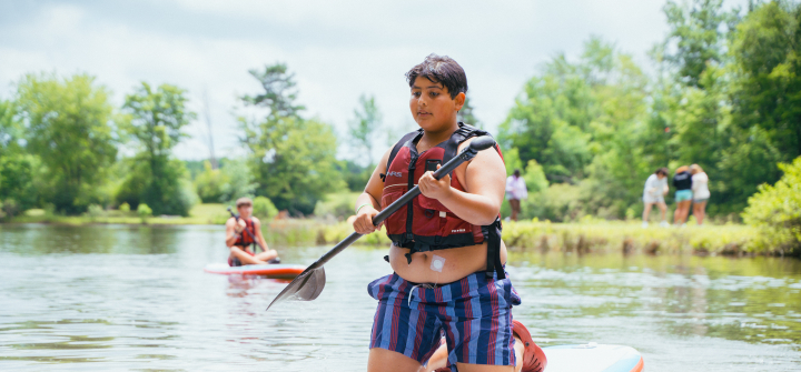 A boy in striped shorts and a life vest, with an insulin patch on his abdomen, paddles a canoe at Camp Ho Mita Koda 