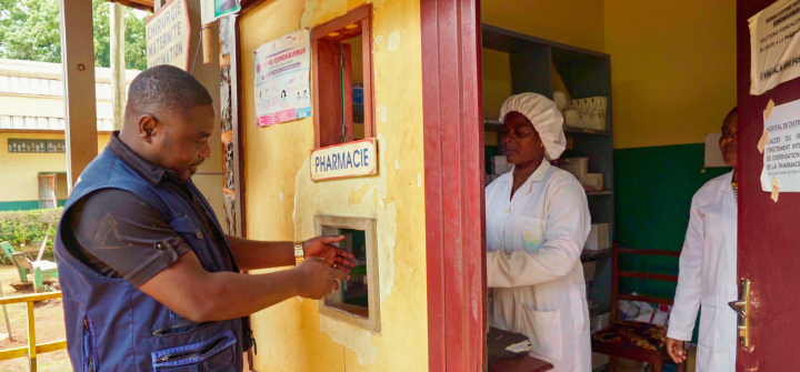 A man wearing a blue vest puts his hands out to receive medicine through a window cut out in a yellow-painted pharmacy.