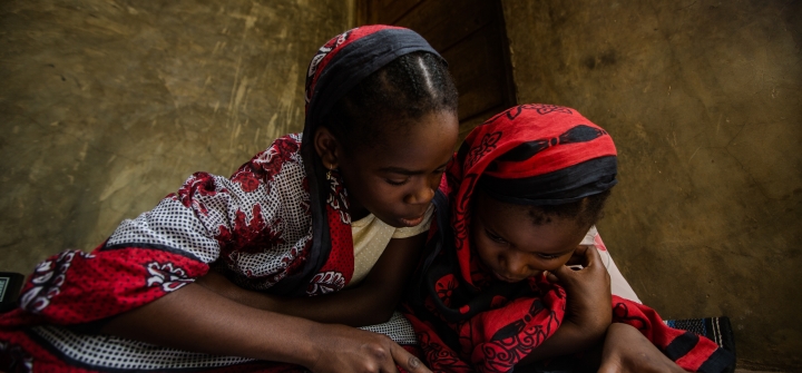Two girls dressed in red and black garments sit on the floor reading USAID malaria educational materials in Zanzibar, Tanzania. 