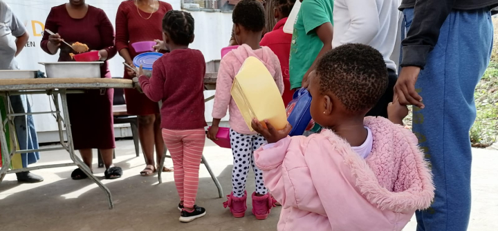Children, including a toddler in a pink coat holding a yellow lunch container, line up to collect a meal. 