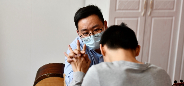 Zhou Pengcheng, wearing a blue surgical mask, clasps hands with his musical therapy patient, a child with short dark hair, shown from the back wearing a long-sleeved grey shirt.