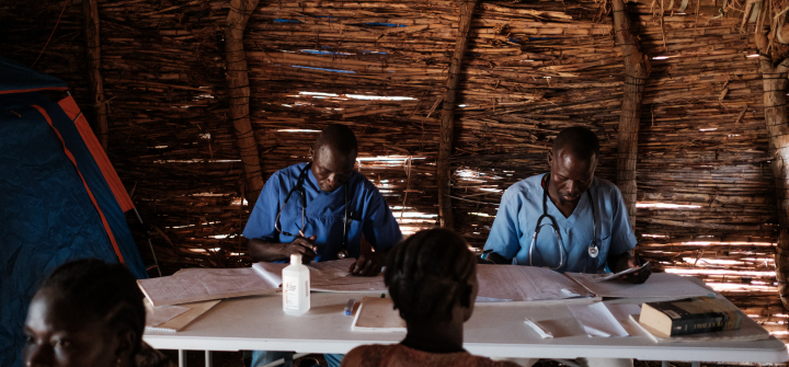 Two doctors wearing blue surgical scrubs seated at a table inside a makeshift wood structure consult with patients standing nearby.