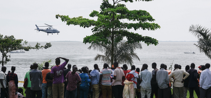 Dozens of bystanders look on as they watch rescuers in a helicopter search the site of a capsized cruise boat on Lake Victoria near Mutima village, south of Kampala, Uganda, on a grey, cloudy day.