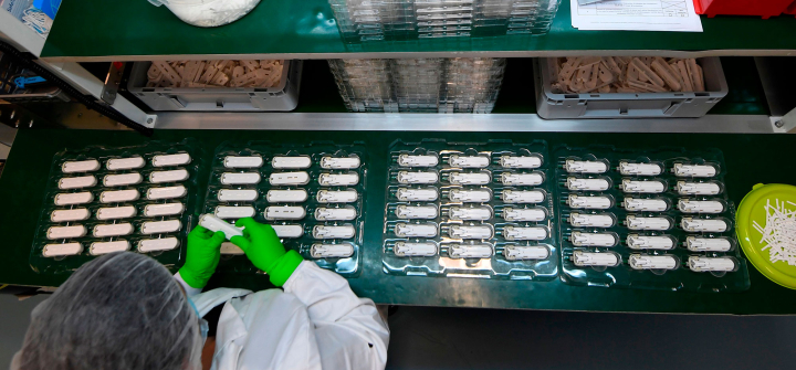 Photo shot from above of an NG Biotech employee wearing green latex gloves, a white coat, and a protective hair covering, working on rows of small white "Carba" antibiotic resistance tests.