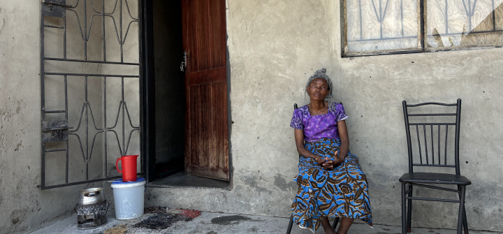 Mary Kapaipi, dressed in a bright purple and blue outfit, sits on the porch of her home with her hands clasped; a red water pitcher and bucket sits next to her door.