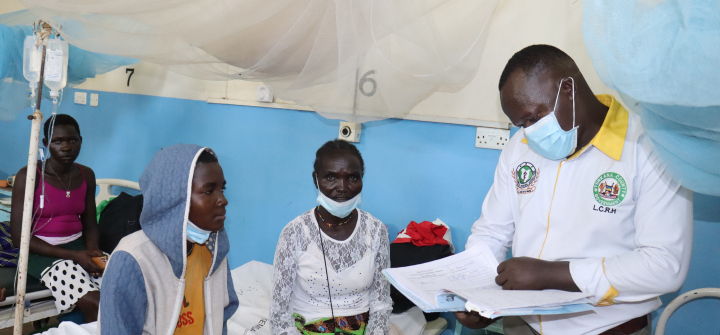 A doctor, standing and looking over a chart, attends to a patient seated on a cot.
