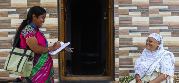 Community health worker Suraiyya Terdale smiles and writes in a notebook as she speaks with a woman wearing a white patterned scarf who is seated with her hands clasped outside a building.