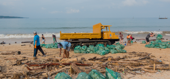 Volunteers remove plastic and other debris from a beach in Kedonganan, Indonesia; dozens of bright green trash bags holding bagged-up debris are piled up around a yellow-orange crawler dump truck.