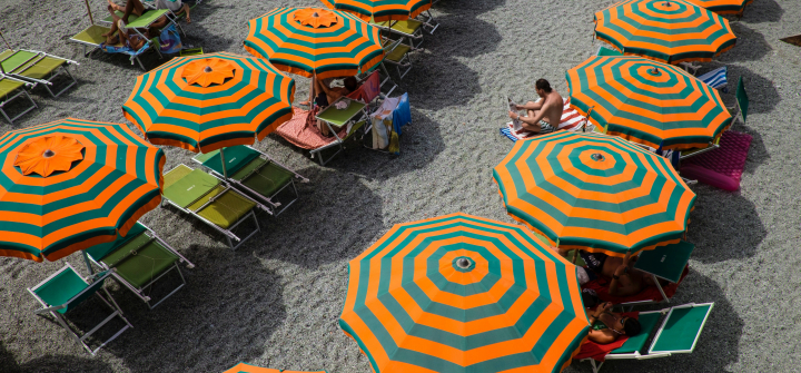Orange and greenish-blue striped beach umbrellas lined up on an Italian beach with a few people sitting underneath.