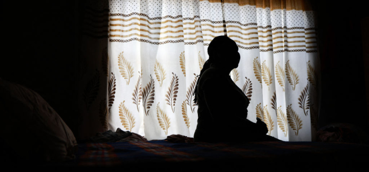 A woman, shown in shadow, sits posing in profile against the backdrop of a light white curtain with beige and brown designs.