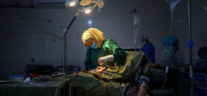 A female surgeon wearing a blue surgical mask and a yellow headscarf operating on a patient.
