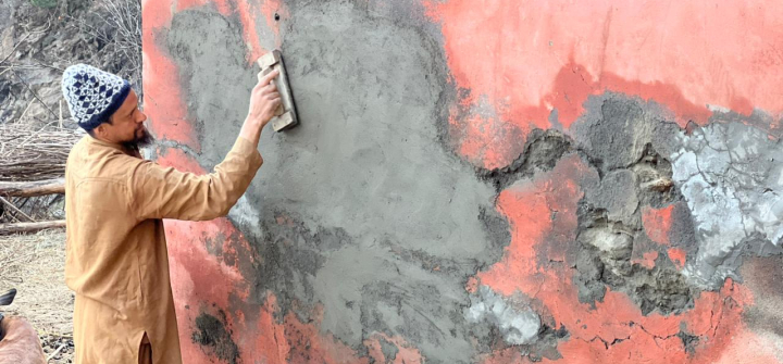 Mohammad Aftab, a man of about 30 years old with a blackish-brown beard, wearing a tan tunic top and pans and a blue and white hat, works applying a grayish, concrete-like mixture to a pink wall. 