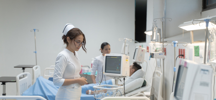 A nurse wearing a white uniform and hat stands next to a patient lying in a hospital bed and wearing a pastel green gown.