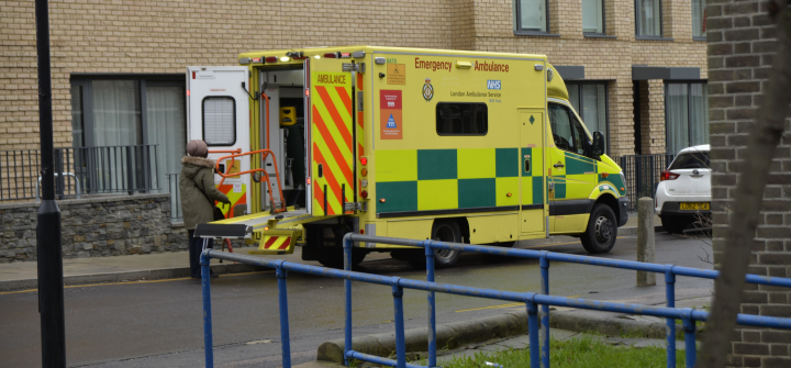 a.	A yellow ambulance with a green checkerboard print on the side sits outside a building as a person wearing a grey coat stands by the open back doors. 
