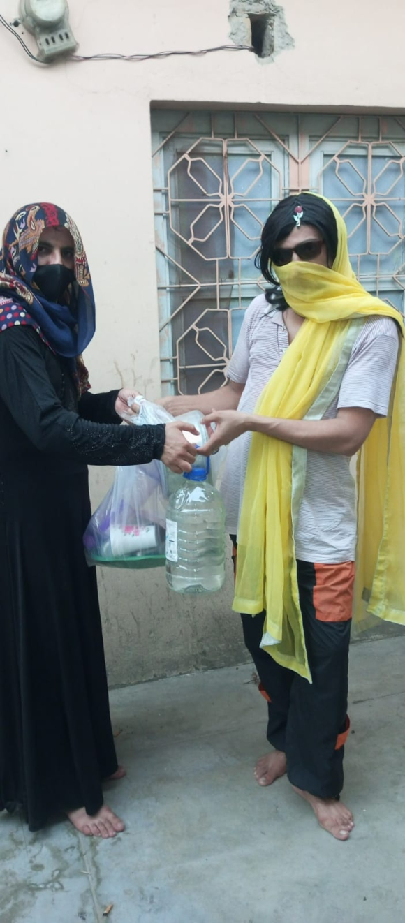 Katrina, a transgender person wearing a black veil, is handing out essential items including drinking water to a friend dressed in a white t-shirt, yellow scarf, and sunglasses.