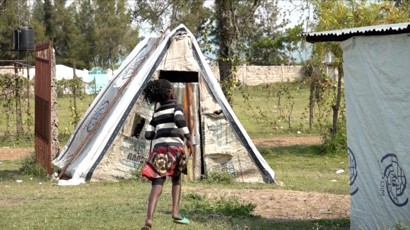 A child wearing a black and white striped long-sleeve shirt and red patterned skirt walks in front of a small tent.