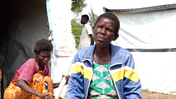 Nduru Camp residents Hellen Anyango wearing a blue and yellow striped jacket and (center) and Serfine Achar, kneeling behind (right) and wearing a red patterned top and orange patterned pants.