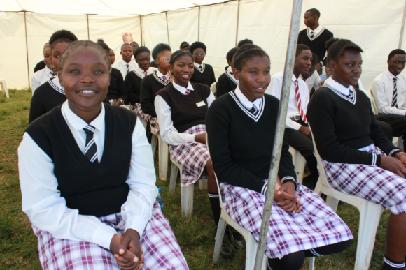 Uniformed schoolgirls sit with their hands clasped in their laps listening to a speaker under a tent.