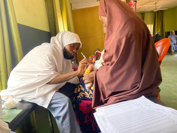 A health worker vaccinates a child held by a mother wearing a rust-colored headscarf  in the Farfaru clinic in nothern Nigeria. 