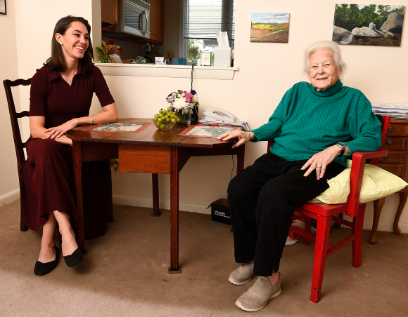 Writer Natalie Draisin in a maroon dress sits at a table with Sue Baker, wearing a green turtleneck and black pants.