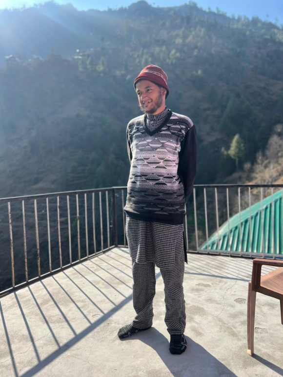 Shabir Ahmed, a 35-year-old man, stands on a balcony with hills in the background, wearing a red knit hat and black and white patterned sweater. 