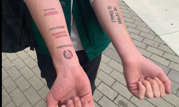 A man displays his arms with tattoos saying "She Decides" and "The Future Is Female" on his forearms.