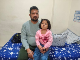 Anil, in green pants and a gray long sleeeved shirt, sitting next to his daughter Nitisha, in a pink long sleeved shirt and jeans, on a bed with a blue blanket with white stars.