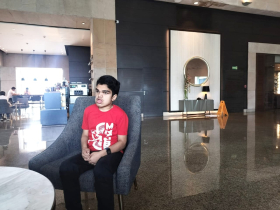 Image of Shaurya Singh, wearing a red shirt with white lettering and black jeans, seasted with his hands in his lap on a gray chair in a lobby with very shiny floors.